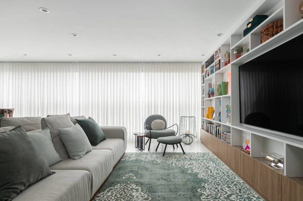 Com base neutra e iluminação abundante, o living integra estar e varanda. A estante sob medida e o tapete em tom esverdeado reforçam a atmosfera acolhedora do ambiente (Foto: Gisele Rampazzo/Divulgação)